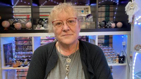 A woman with curly blonde hair tied back. She has red framed glasses and is wearing a grey t-shirt and black cardigan and several silver necklaces with gems and crystals. Behind her are shelves lit up in soft purple light with crystals and precious stones on them