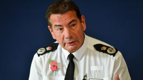 Nick Adderley, wearing a white police shirt with black tie and police badges on his epaulettes. He is also wearing a poppy on his chest and has a pen in his breast pocket. He is sitting in front of a blue background. 