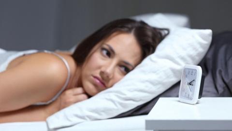 A woman with dark hair lying on a pillow starring at an alarm clock
