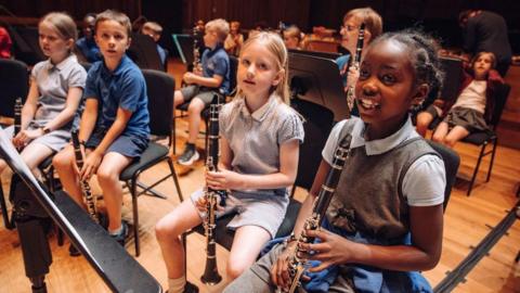 Children from Horfield CE Primary school at Earthsong day at Bristol Beacon, 2024. The children are in school uniform style outfits and the main children - four of them, are all holding clarinets. They have music stands in front of them and are all sitting as if ready to begin playing. 