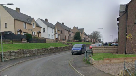 A road going up a hill with houses on either side. The houses on the left side are raised on a hill with grass in front of them and bordered by a stone wall. Cars are parked along the side of the road.