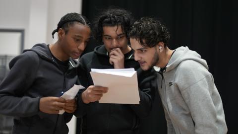 Three young men anxiously read the one in the middle's A-level results. He is holding his hand up to his mouth. 