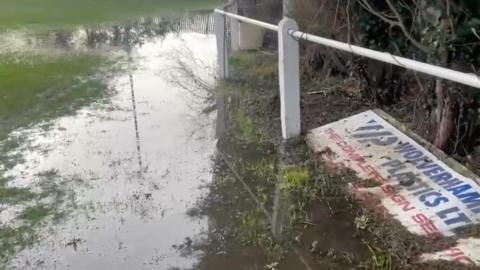 A heavily waterlogged area beside a football pitch with standing water submerging the grass. A white perimeter railing and a partially visible advertising board are lying on the ground.