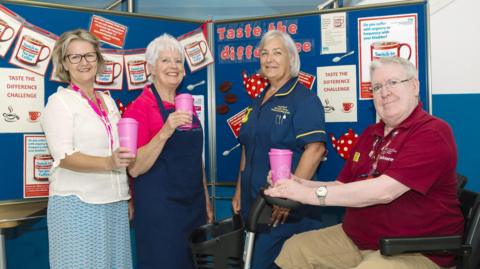 Three women are standing in front of a blue bulletin board with posters all over it. A sign on it reads "taste the difference". They are holding pink reusable cups. There is a man on the right hand side sitting in a motorised scooter, also holding a pink reusable cup