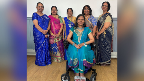 A group of 6 Hindu women dresses in sarees with mostly different shades of blue. They have on jewellery and one of them is sitting in a wheelchair. They are wearing a selection of fabric's mainly in different hues of blue and shiny materials. They are smiling and  have a beautiful selection of women's Indian sarees.