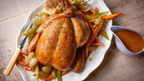 A whole roast chicken sitting on a bed of vegetables on a large white plate. A gravy boat is next to it.