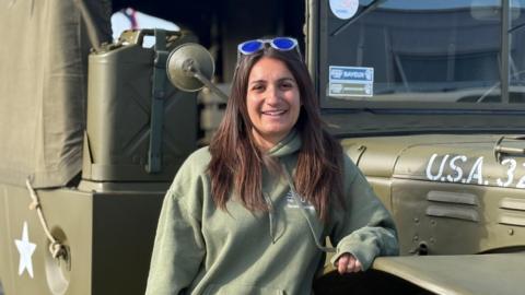 A woman in her early 30s with long brunette hair. She is wearing a military green jumper and is stood in front of a World War Two military truck.