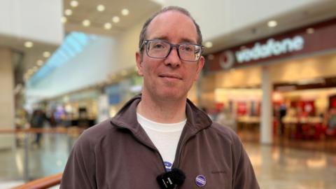 A head-and-shoulders photo of a man with short grey hair, wearing glasses and a brown fleece, looking straight at the camera. He's inside a shopping centre and there's a row of shops behind him.