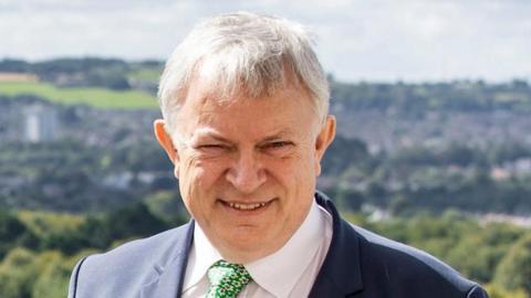 A man with grey hair smiling at the camera. Witha navy suit, white shirt and green tie. In the background there are faded green country fields 