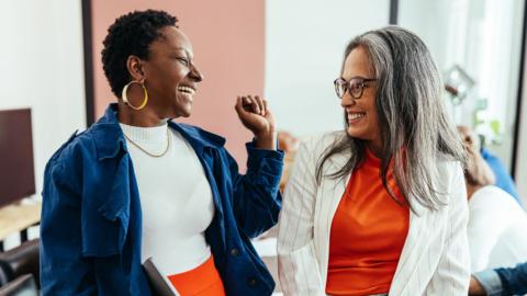 Two women laugh at each other in an office setting.