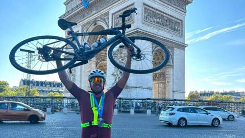 Man holding bicycle aloft in front of the Arc de Triomphe after completing what some call the world's toughest triathlon