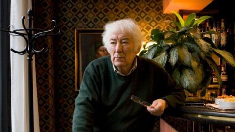 heaney sits on a bar stool. Behind him is a bar with a plant, and bottles on the shelves and nuts on the corner. He looks away off camera. 