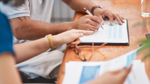 Two people are filling out a form on a wooden surface. The person furthest away is holding a silver pen and is writing on the form. The person closest is pointing towards the form. There is a pair of glasses and a glass also on the surface. The closest person is holding paperwork also. 