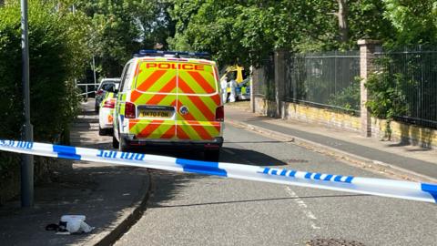Police vehicles parked on a road behind a police cordon. The road is cordoned off with blue and white police tape. People in white suits can be seen in the distance.