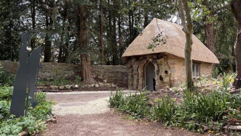 A small stone building with a thatched roof, a window, and a door. It resembles a fancy hut and is in a garden surrounded by trees and a stone wall.