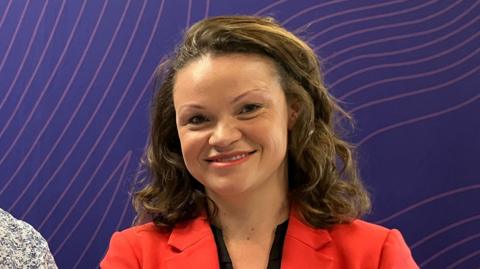 Council leader Bella Sankey has long brown wavy hair and wears a bright red suit jacket over a black top. This photo is taken in a radio studio with a purple background.