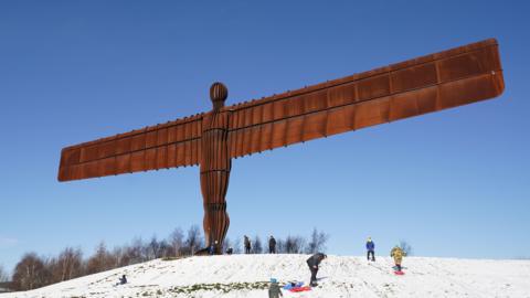 Photo of the Angel of the North in the snow with people sledding beneath it. The Angel of the North is a brown sculpture of an angel with very long wings. 