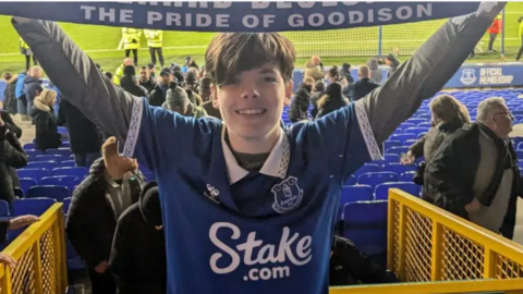 A smiling Mackenzie holds an Everton scarf over his head with the Goodison Park pitch behind him