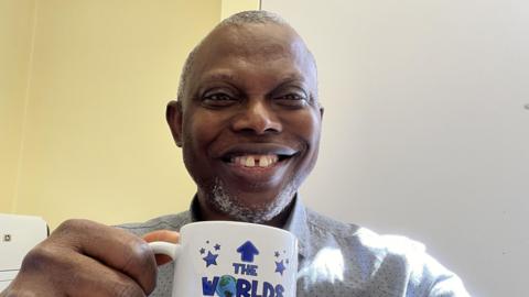 Dumbor Ngaage smiles at the camera as he holds a mug that reads "the world's best professor". He is wearing a white shirt covered in tiny dots. He has a short cropped beard and short grey hair. 