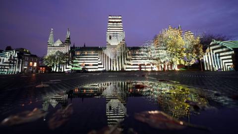 'Liquid Geomotry' lights up Durham Cathedral during a preview for Lumiere 2023. The sprawling  medieval stone building is illuminated in black and white vertical and horizontal stripes, reminiscent of a geometric zebra. The building and lights are reflected in water in the foreground.
