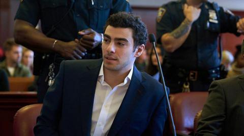 Luigi Mangione, wearing a blazer and white button-down, sitting in front of a microphone in court