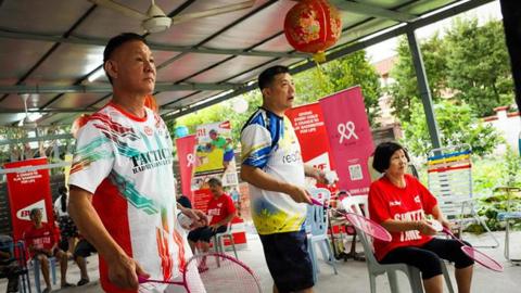 Two men stand up and a woman sits down, with all three holding badminton racquets.
