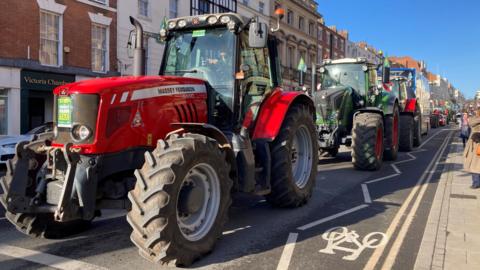 Three tractors drive down a high street on a sunny day. The tractor at the front is red, the second is green, and the third is red. A traffic queue can be seen behind the tractors.