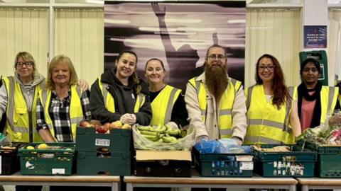 Some of the volunteers at Helping Hearts, all wearing yellow hi-vis vests, stand behind a row of tables containing green plastic baskets full of food
