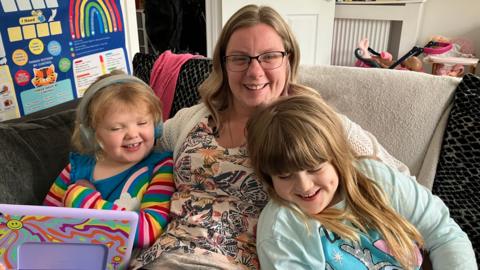 Hayley sitting on the sofa in her living room with her arms around her two daughters. They are all smiling and playing, wearing colourful clothes. Hayley has long way strawberry blond hair and glasses and is wearing a beige blouse with butterflies on it.