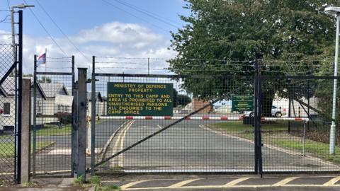 Black gates which lead to an army base. There is a large green sign with yellow writing, telling people entry is prohibited.