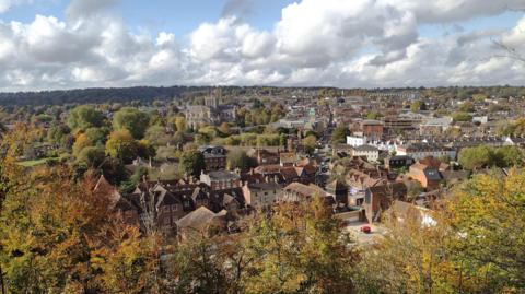 An aerial view over the centre of Winchester. In the centre of the frame you can see the large stone cathedral. The city is made up of stone and brick buildings and surrounded by trees that are turning shades of orange and brown.