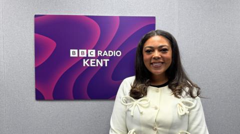An image of Tia Davies stood in front of the BBC Radio Kent logo. Tia is wearing a white jacket with bows. 