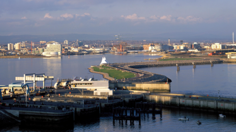 Llun o forglawdd Bae Caerdydd o'r awyr ar ddiwrnod braf gyda'r brifddinas yn y cefndir