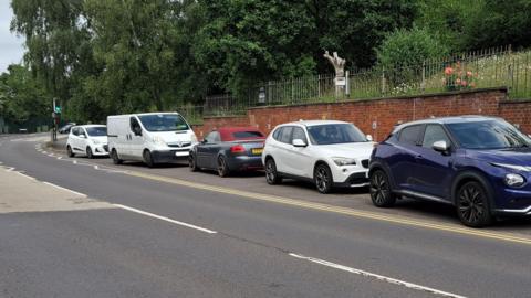 Cars and a van of various colours parked on a layby just past the bend of a road. Behind the cars is a red-brick wall topped with iron railings. Behind the railings is a grassy bank topped with green trees.
