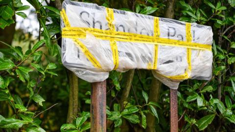 A sign wrapped in plastic sits in front of green foliage. It's wrapped in yellow tape. The two legs of the sign are rusted. There are brown leaves on the ground.  