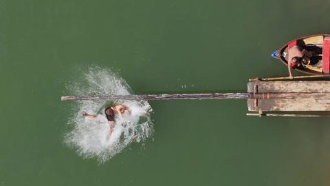 An aerial view of a man in black trunks falling into green seawater, making a large splash. He has fallen from a beam that is stretched out over the water from a jetty. There is a man in a boat to the right of the image, who is wearing red trunks. 