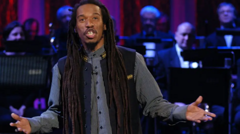 A man with long dreadlocks, wearing a blue patterned shirt and a blue waistcoat embroidered in gold, holds his hands out as he speaks. A black tie orchestra is behind him.