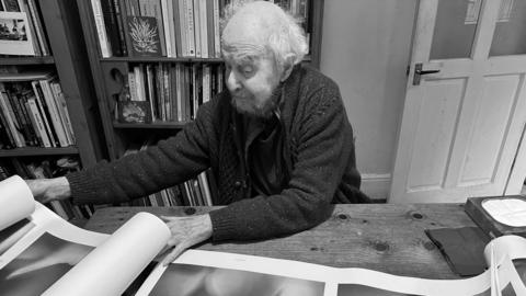 An image of John sat at a wooden table rolling out a series of prints. He had straggly light hair around a balding head, a beard and wears a cardigan while sitting at the table. Behind him are bookshelves with dozens of books on and an open door.