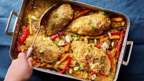 A large metal tray filled with katsu-style chicken traybake, a hand holding a large metal spoon is digging in,
