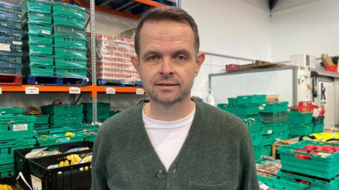 Nathan Atkinson stands looking at the camera. He is wearing a green cardigan over a white t-shirt. He has short brown hair. Behind him are a number of green a black boxes stacked on top of each other. The boxes contain fresh fruit and vegetables.