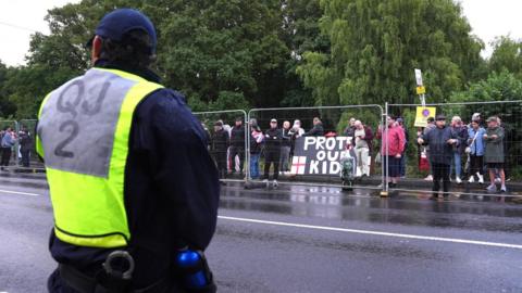 The back of a uniformed police officer, who is wearing a high-vis yellow jacket. He is looking across a road, and on the other side is a metal fence and crowds gathered, including a banner that reads "protect our kids".