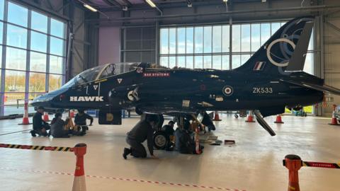 Two groups of production workers check the components underneath a dark-coloured BAE Hawk jet at a Lancashire base