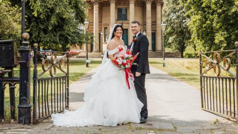A woman in a white wedding dress stands with a man in a suit. She holds a bouquet of red and pink flowers with a red ribbon. They stand at the pathway to a round church, metal ornate gates are open next to them and the path lined with traditional street lights. To the right and left is a lawn with large trees.