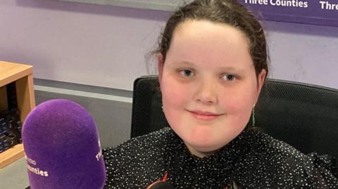 Betsy Griffin is in the BBC 3CR radio studio sitting in a chair in front of the purple microphone. She is smiling at the camera and is wearing a sparkly black top.