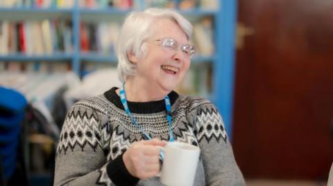A lady laughs as she holds a tea mug. She has grey hair and is wearing glasses and a grey patterned jumper.