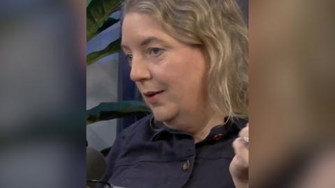 A woman with wavy hair and a dark coloured shirt looks at someone off camera