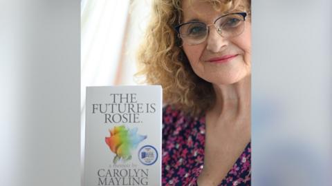 Carolyn Mayling stood holding her book and looking to the camera