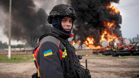 A member of the emergency services in Sumy, dressed in high vis with the Ukraine flag sewn onto his arm and a combat helmet and balaclava, looks wearily at the camera while standing in front of a burning gas depot near the Russian border.