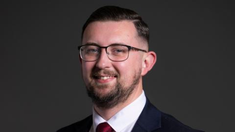 Connor Naismith, a man wearing glasses and a blue suit with white shirt and red tie, is smiling at the camera. He is standing against a dark background.