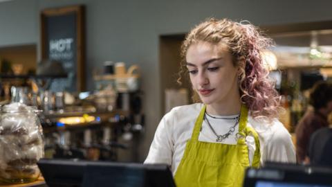 A young woman wearing a green apron looks at a till in a cafe.